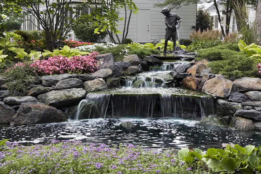 Closer to the home, a pond and waterfall showcase the family’s beloved bronze statue. Surrounded by shrubs and perennials, the outdoor art personalizes the landscape and makes it fully unique. The bronze figure was sculpted from a photograph of the homeowner as a child. Water pours from his bucket onto the stone and into the pond. Due to its location at the property’s low point, pond drainage presented a challenge. French drains were installed to catch water flowing from the house, drive and lawns to prevent overflow.